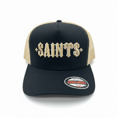 Black and oat MUKA Saints trucker hat with mesh back and curved brim, casual accessories for a stylish, laid-back look.